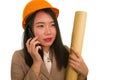 Beautiful and happy Asian Korean woman in hardhat holding building blueprints as architect engineer or constructor supervisor Royalty Free Stock Photo