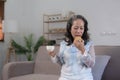 Beautiful and happy Asian 60s aged woman having breakfast, sipping a cup of tea and eating cookies in her living room. Royalty Free Stock Photo