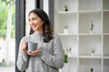 Beautiful Asian woman in cozy sweater holding a cup of coffee, smiling, looking out the window Royalty Free Stock Photo