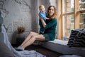 Beautiful happy blue-eyed baby sitting with her mother on the windowsill. They look at the camera and show their language Royalty Free Stock Photo