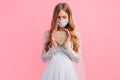 Bride in a wedding dress with a medical protective mask on her face, holding a wooden heart on a pink background. Quarantine, Royalty Free Stock Photo