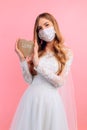 Bride in a wedding dress with a medical protective mask on her face, holding a wooden heart on a pink background. Quarantine, Royalty Free Stock Photo
