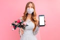 Bride in a wedding dress with a medical protective mask on her face, shows a blank mobile phone screen on a pink background. Royalty Free Stock Photo