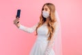 Bride in a wedding dress with a medical protective mask on her face, taking a selfie on a smartphone on a pink background.