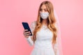 Bride in a wedding dress with a medical protective mask on her face, using a mobile phone on a pink background. Quarantine, Royalty Free Stock Photo