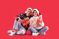 Beautiful happy couple in Santa hats and sweaters sitting with Christmas gifts on red background Royalty Free Stock Photo