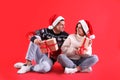 Beautiful happy couple in Santa hats and sweaters sitting with Christmas gifts on red background Royalty Free Stock Photo