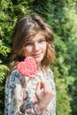 Beautiful happy girl with a big red candy on a stick, smiling and laughing. Summer tree. Green bush.