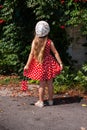 Beautiful happy girl in red polka dot dress on green leaves background.Cute joyful child with long blonde hair back view