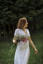 A beautiful happy girl in a white dress holds a bouquet of wild flowers in her hands and turns around. Portrait of a slender girl