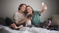 Beautiful happy lesbian couple sitting in pajamas on the bed in the morning making selfie on smartphone with dog bassengi Royalty Free Stock Photo