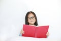 Beautiful happy little Asian kid girl reading hardcover book lying on bed against white background Royalty Free Stock Photo