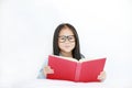 Beautiful happy little Asian kid girl reading hardcover book lying on bed against white background Royalty Free Stock Photo