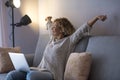 Beautiful happy mature woman in curly hair with arms outstretched and eyes closed sitting with laptop on sofa at home. Satisfied