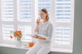 Beautiful happy middle age woman sitting on windowsill at home and reading book. Young woman with short hair eating macaroons and Royalty Free Stock Photo