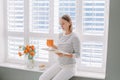 Beautiful happy middle age woman sitting on windowsill at home and reading book. Young woman with short hair drinking tea coffee Royalty Free Stock Photo