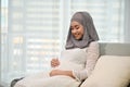 Happy Asian Muslim pregnant woman touching her belly, sitting on her couch in the living room Royalty Free Stock Photo