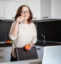 Beautiful happy modern charming elderly woman in glasses in the kitchen with a glass of water and an orange in her hands, the Royalty Free Stock Photo