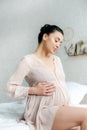 Happy pregnant woman in nightie touching tummy while sitting on bed Royalty Free Stock Photo