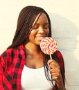 Beautiful happy smiling african woman is enjoying sweet lollipop, wearing a red checkered shirt in the city Royalty Free Stock Photo