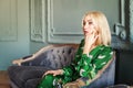 Beautiful happy smiling blonde woman in green dress sitting on velvet sofa Royalty Free Stock Photo