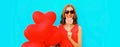 Beautiful happy smiling young woman with lollipop and bunch of red heart shaped balloons wearing sunglasses on blue background Royalty Free Stock Photo