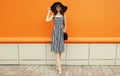 Beautiful happy smiling young woman posing full-length wearing black summer straw hat, striped dress with handbag on orange Royalty Free Stock Photo