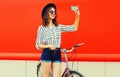 Beautiful happy smiling young woman taking selfie by smartphone with bicycle wearing a shirt, shorts, black round hat on red Royalty Free Stock Photo