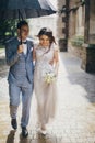 Beautiful happy wedding couple embracing together under umbrella in sunny rainy street. Stylish bride and groom walking and Royalty Free Stock Photo