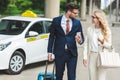 beautiful happy young couple in formal wear smiling each other while going with suitcase near taxi