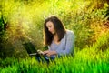 Beautiful happy young girl sits in a park on a glade with a laptop in green grass. sunny day Royalty Free Stock Photo