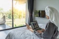 Beautiful happy young woman in a gray bathrobe and with a towel on her head on a bed in a hotel room working on a laptop, vacation Royalty Free Stock Photo