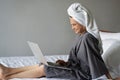 Beautiful happy young woman in a gray bathrobe and with a towel on her head on a bed in a hotel room working on a laptop, vacation Royalty Free Stock Photo