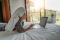 Beautiful happy young woman in a gray bathrobe and with a towel on her head on a bed in a hotel room working on a laptop, vacation Royalty Free Stock Photo