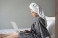 Beautiful happy young woman in a gray bathrobe and with a towel on her head on a bed in a hotel room working on a laptop, vacation Royalty Free Stock Photo