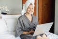 Beautiful happy young woman in a gray bathrobe and with a towel on her head on a bed in a hotel room working on a laptop, vacation Royalty Free Stock Photo