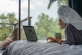 Beautiful happy young woman in a gray bathrobe and with a towel on her head on a bed in a hotel room working on a laptop, vacation Royalty Free Stock Photo