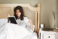 Beautiful happy young woman reading book and drinking coffee in bed in hotel room or home bedroom. Stylish brunette girl in white Royalty Free Stock Photo