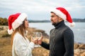 Beautiful heterosexual couple in love toasting outdoor wearing Santa Claus hat looking into each other`s eyes holding white wine