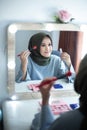 Beautiful hijab woman is dressing up in front of the mirror while holding brush and lipstick with smile expression. Hijab girl at Royalty Free Stock Photo