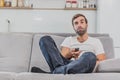 Beautiful hilarious young man holding a remote control. During this, the TV is watching while sitting on the couch at