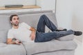 Beautiful hilarious young man holding a remote control. During this, the TV is watching while sitting on the couch at