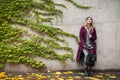 A Beautiful Hipster Girl In A Purple Coat On A Concrete Gray Wall Background And Green And Yellow Autumn Leaves. Space For Text