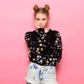 Beautiful hipster thinking girl with pigtails wearing funny sweater and jeans posing on pink background.