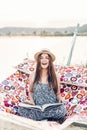 Beautiful hipster woman relaxing in a hammock, emotional girl in stylish hat reading book while resting on the beach at sunset Royalty Free Stock Photo