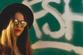 The hipster young girl wearing fashionable sunglasses and black hat with a graffiti as background