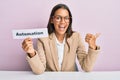 Beautiful hispanic business woman holding automation word on paper pointing thumb up to the side smiling happy with open mouth Royalty Free Stock Photo