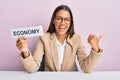 Beautiful hispanic business woman holding economy message paper pointing thumb up to the side smiling happy with open mouth