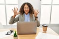 Beautiful hispanic business woman sitting on desk at office working with laptop showing and pointing up with fingers number nine