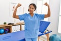 Beautiful hispanic physiotherapist woman at pain recovery clinic showing arms muscles smiling proud Royalty Free Stock Photo